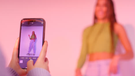 Studio-Shot-Of-Woman-Taking-Photo-Of-Friend-Dancing-On-Mobile-Phone-Against-Pink-Background-2